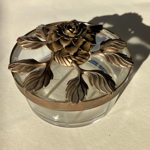 Pode incluir: Um frasco de vidro transparente com uma tampa de metal cor de bronze. A tampa é decorada com uma rosa e folhas. O frasco tem uma faixa de metal cor de bronze ao redor do meio.