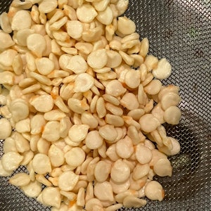 May include: A close-up shot of a pile of off-white pepper seeds. The seeds are small, flat, and oval-shaped, with a slightly translucent appearance. They are resting in a fine mesh strainer, which is partially visible in the background.