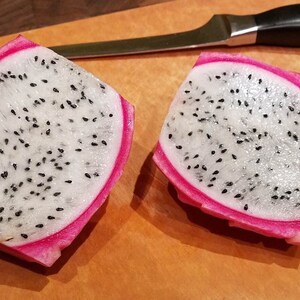 May include: Two halves of a dragon fruit, cut open to show white flesh with black seeds. The fruit has a bright pink skin. A knife with a black handle is on a wooden chopping board.