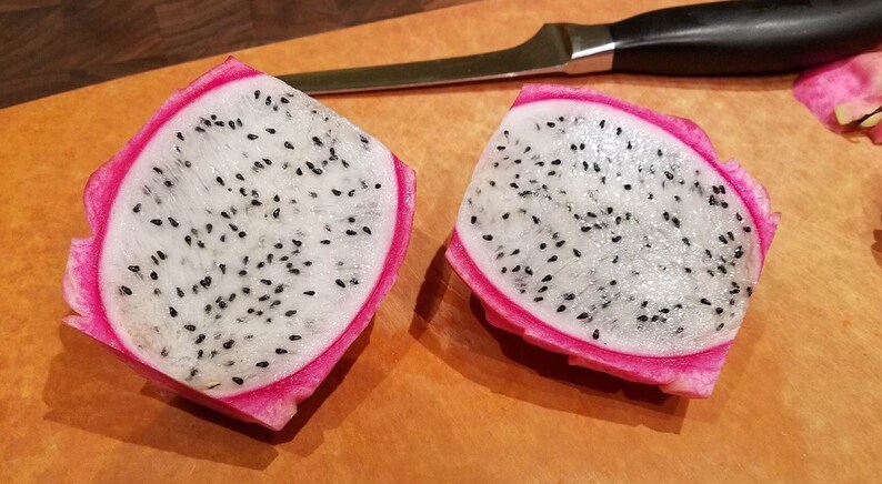 May include: Two halves of a dragon fruit, cut open to show white flesh with black seeds. The fruit has a bright pink skin. A knife with a black handle is on a wooden chopping board.