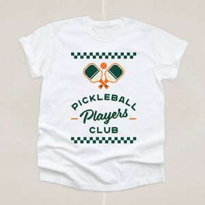 May include: White t-shirt featuring a green and orange pickleball paddle graphic with the text "Pickleball Players Club" in a black checkered border.