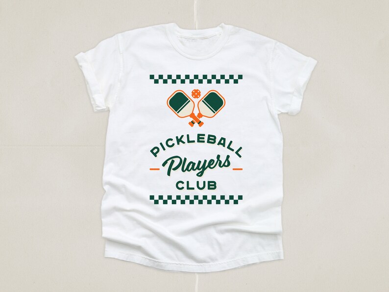 May include: White t-shirt featuring a green and orange pickleball paddle graphic with the text "Pickleball Players Club" in a black checkered border.