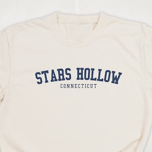 May include: A cream-coloured t-shirt featuring the navy blue text "STARS HOLLOW CONNECTICUT" arched across the chest. The shirt has a crew neck and short sleeves, with the sleeves rolled up.