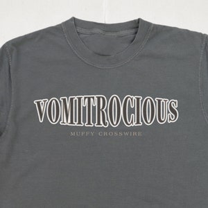 May include: A dark grey t-shirt with the text "VOMITROCIOUS" in a white outline with black fill. The text "MUFFY CROSSWIRE" is printed below in a smaller font.