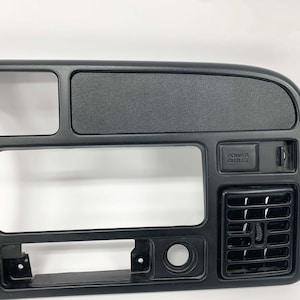 Op de afbeelding: Zwarte kunststof dashboardbekleding met een stopcontact en een ventilatieopening. Het paneel heeft een rechthoekige uitsparing voor een radio en een kleine rechthoekige uitsparing voor een opbergvak.