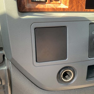 May include: A close-up of a gray car interior with a black rectangular panel and a cigarette lighter.