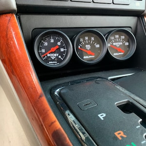 May include: Three black gauge pods with white faces and red needles. The gauges are labeled "Boost", "Oil", and "Water". The gauges are mounted in a car's center console.