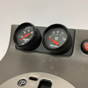May include: Two black gauges mounted in a grey dashboard. The left gauge is labelled "BOOST" and reads 20 PSI. The right gauge is labelled "OIL PRESS" and reads 60 PSI. The gauges are made by Auto Meter.