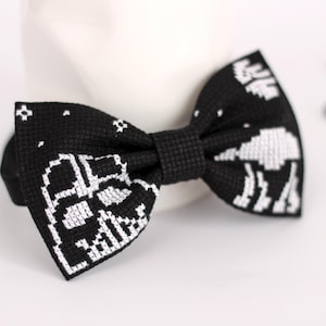 May include: A black bow tie with a white pixelated design of a spaceship. The bow tie is made of a soft, textured fabric.