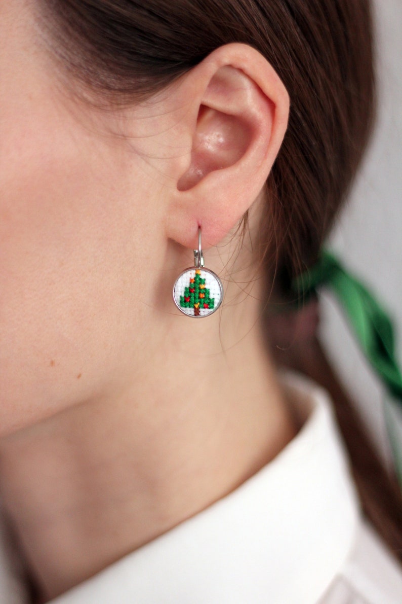 Op de afbeelding: Een paar zilveren oorhangers met een kleine ronde witte hanger met een groene kerstboom met gele en rode ornamenten.