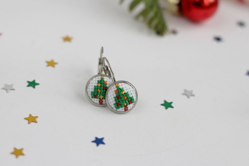 Op de afbeelding: Een paar zilveren oorbellen met een witte achtergrond en een groen kerstboomontwerp. De boom is versierd met rode en gele kerstballen.
