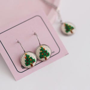 Op de afbeelding: Een paar zilveren creolen met een witte achtergrond en een groen geborduurd kerstboomontwerp. De oorbellen worden gepresenteerd op een roze kaart.