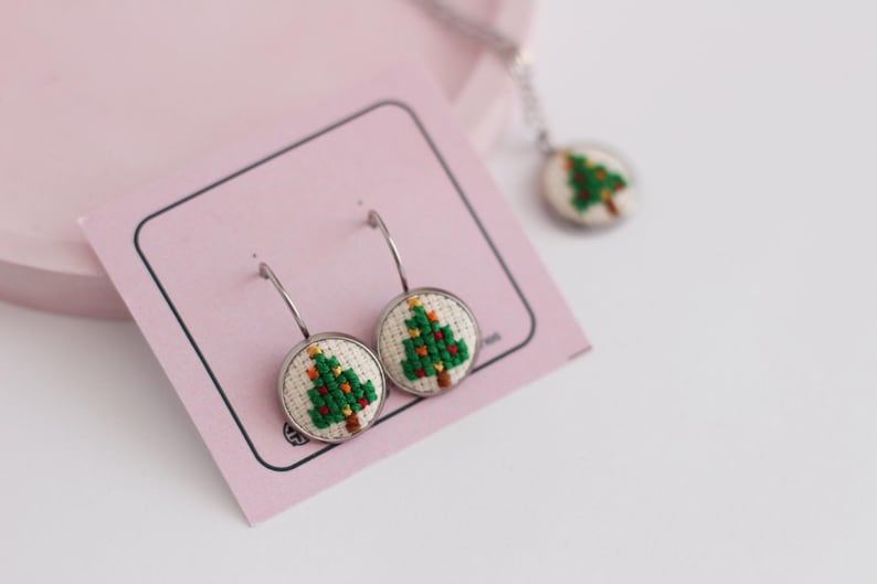 Op de afbeelding: Een paar zilveren creolen met een witte achtergrond en een groen geborduurd kerstboomontwerp. De oorbellen worden gepresenteerd op een roze kaart.