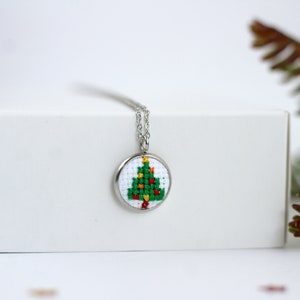 Op de afbeelding: Een zilveren ketting met een ronde hanger met een groene kerstboom met rode en gele ornamenten, gekruist gestikt op witte stof.