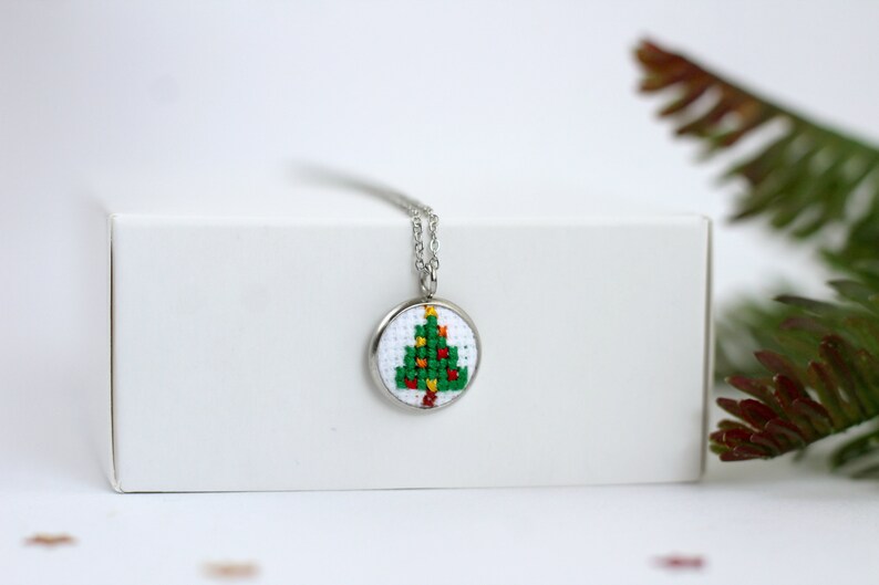 Op de afbeelding: Een zilveren ketting met een ronde hanger met een groene kerstboom met rode en gele ornamenten, gekruist gestikt op witte stof.