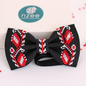 May include: A black bow tie with a red and white embroidered floral pattern. The bow tie has a black adjustable strap. The brand name "nzee authentic accessories" is visible on a teal oval in the background.