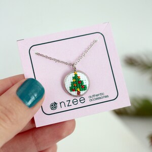 Op de afbeelding: Een zilveren ketting met een klein rond hanger met een geborduurd kerstboomontwerp in groen en rood. De hanger bevindt zich op een roze kaart met de tekst "nzzee authentic accessories".