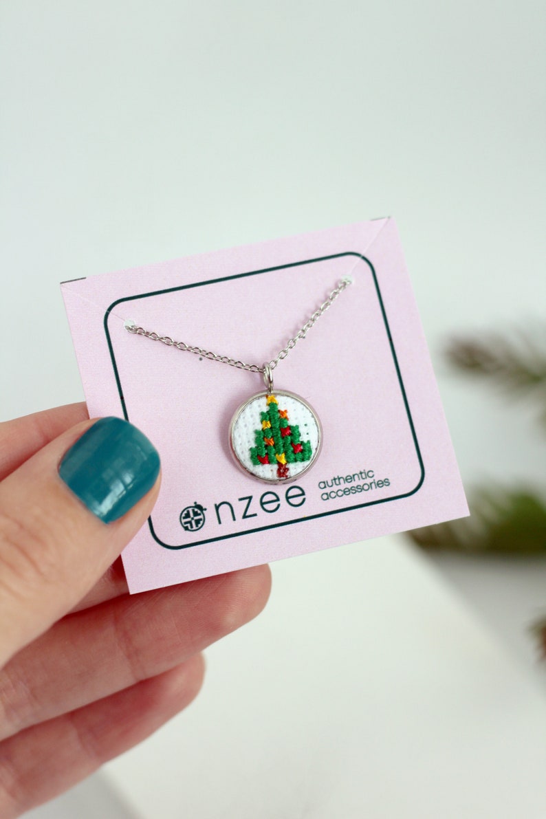 Op de afbeelding: Een zilveren ketting met een klein rond hanger met een geborduurd kerstboomontwerp in groen en rood. De hanger bevindt zich op een roze kaart met de tekst "nzzee authentic accessories".