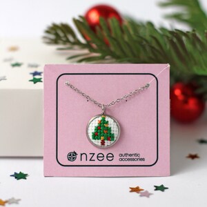 Op de afbeelding: Een zilveren ketting met een kleine ronde hanger met een groen en rood kerstboom-kruissteekontwerp. De hanger bevindt zich op een roze kaart met de tekst "nz&eacute;e authentic accessories".