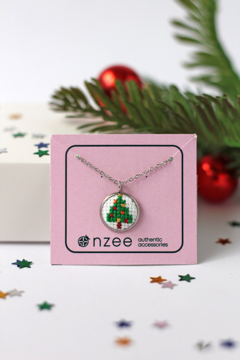 Op de afbeelding: Een zilveren ketting met een kleine ronde hanger met een groen en rood kerstboom-kruissteekontwerp. De hanger bevindt zich op een roze kaart met de tekst "nz&eacute;e authentic accessories".