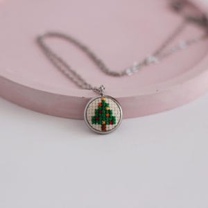 Op de afbeelding: Een zilveren ketting met een ronde hanger met een gestikt kerstboommotief in groen en bruin.