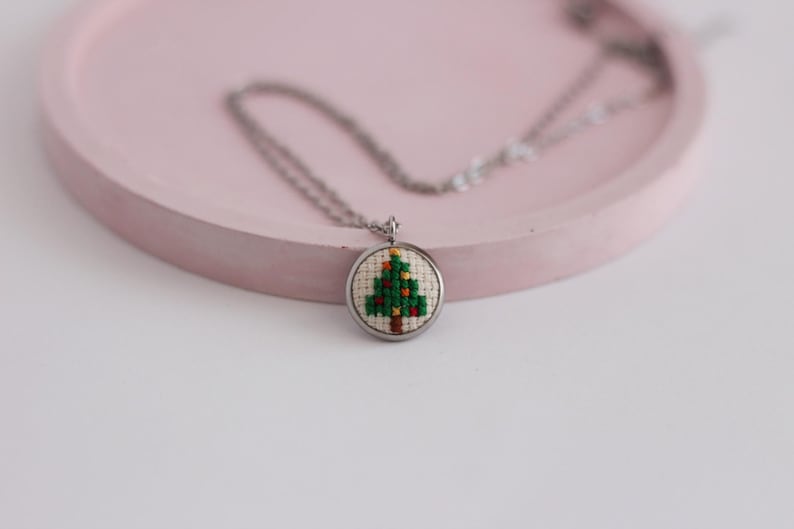 Op de afbeelding: Een zilveren ketting met een ronde hanger met een gestikt kerstboommotief in groen en bruin.