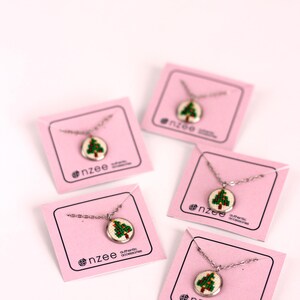 Op de afbeelding: Vier roze kaarten met zilveren kettingen en kleine ronde hangers met een groen geborduurd kerstboomontwerp. Elke kaart heeft de tekst "nzee authentic accessories".
