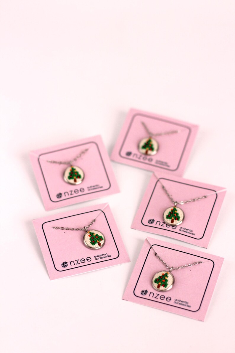 Op de afbeelding: Vier roze kaarten met zilveren kettingen en kleine ronde hangers met een groen geborduurd kerstboomontwerp. Elke kaart heeft de tekst "nzee authentic accessories".