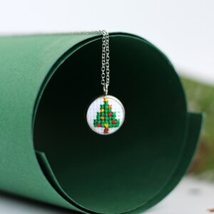 Op de afbeelding: Een zilveren ketting met een klein rond hanger met een geborduurd kerstboomontwerp in groen en rood.