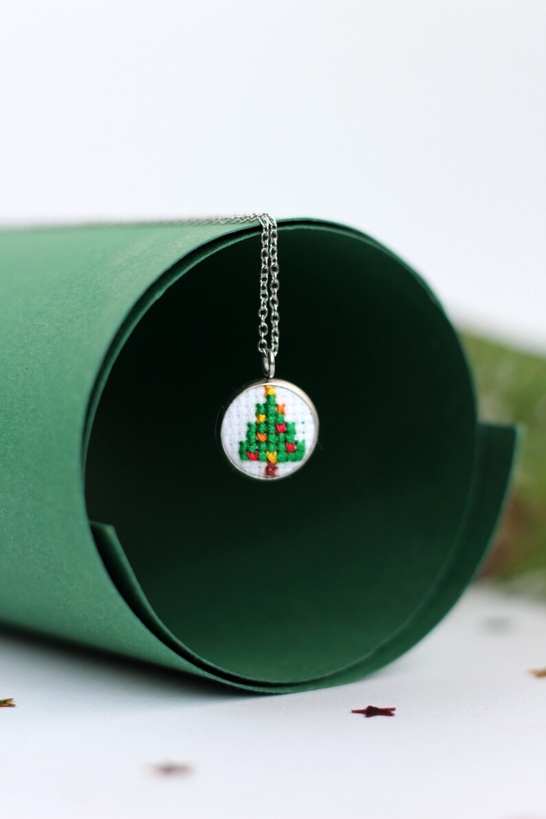 Op de afbeelding: Een zilveren ketting met een klein rond hanger met een geborduurd kerstboomontwerp in groen en rood.