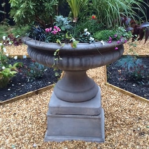 May include: A large, ornate stone planter filled with a variety of plants and flowers. The gray planter sits on a pedestal base. The plants include tall grasses, leafy greens, and small pink flowers, creating a lush garden display.