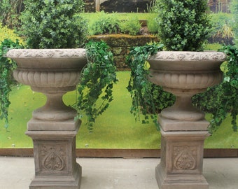May include: Two large, brown stone urns with decorative details, each sitting on a square pedestal. The urns are filled with green leafy plants and are set against a backdrop of green grass and foliage.