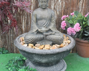 May include: A gray stone fountain with a meditating Buddha statue in the center. The fountain is filled with small, smooth stones and is surrounded by green foliage.