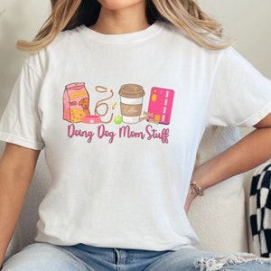 以下が含まれることがあります： 白いTシャツに、ピンクのグラフィックデザインが施されています。デザインには、「Doing Dog Mom Stuff」という文字と、犬のフードバッグ、リード、犬の骨、コーヒーカップ、クレジットカードが描かれています。
