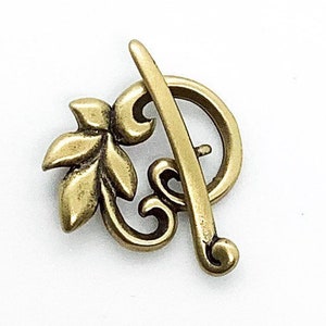 May include: A gold-toned metal clasp with a decorative leaf design. The clasp is shaped like a circle with a leaf on one side and a curved bar on the other.
