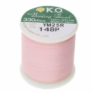Pode incluir: Um carretel de fio para miçangas K.O. rosa claro, 100% nylon, com o texto "YM25R 14BP" e "Made in Japan" impresso na etiqueta. O carretel está sobre uma base branca e tem um rótulo verde.