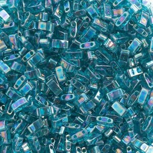 Peut inclure: Gros plan sur un tas de perles rectangulaires bleu sarcelle iridescentes. Les petites perles ont une forme rectangulaire avec deux trous à chaque extrémité. La surface brillante des perles reflète la lumière, créant un effet chatoyant.