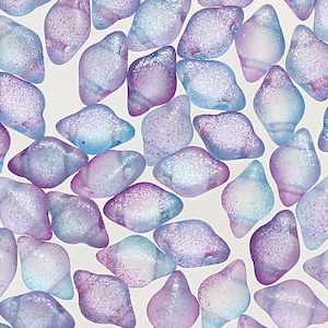 May include: A collection of iridescent, sea glass-like beads in shades of blue, purple, and pink. The beads have a textured, frosted appearance and are scattered across a white background. The beads are irregular in shape and have a glittery finish.