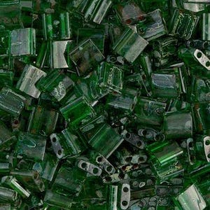 Puede incluir: Primer plano de un montón de cuentas de vidrio verde oscuro. Las cuentas son rectangulares con dos agujeros en cada extremo. Las cuentas tienen un aspecto ligeramente translúcido y la luz se refleja en sus superficies.