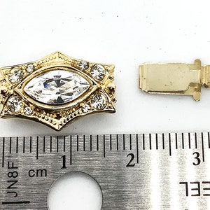 May include: Gold-tone clasp with a large clear marquise-shaped rhinestone surrounded by smaller rhinestones. The clasp measures approximately 1 inch in length.