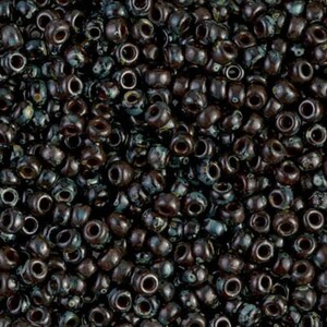May include: A close-up of a collection of dark brown seed beads. The beads have a subtle iridescent sheen, with touches of teal and green. Each bead is pierced with a central hole, and they are randomly arranged.