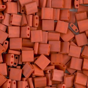 Può includere: Primo piano di un mucchio di perline quadrate color corallo. Ogni perlina ha due fori per l'infilatura. Le perline sono sparse, mostrando varie angolazioni e orientamenti. Le perline hanno una finitura opaca.
