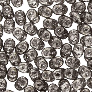 May include: A close-up shot of numerous dark gray, oval-shaped beads with two holes each. The beads are scattered across a white background, creating a textured, organic pattern. Ideal for jewelry making and crafting.