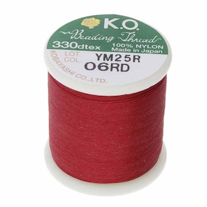 Pode incluir: Um carretel de fio de miçangas K.O. vermelho. O rótulo diz "330dtex 100% NYLON Made in Japan". O carretel é branco com um rótulo verde e dourado. O fio é enrolado firmemente ao redor do carretel.