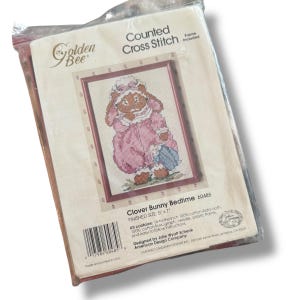 May include: A cross-stitch kit featuring a framed image of a bunny in a pink nightgown holding a blue umbrella. The kit includes Aida cloth, cotton floss, a needle, and a plastic frame. The finished size is 5" x 7". The packaging is clear plastic.