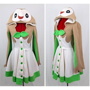Puede incluir: Un vestido blanco y marrón con capucha y detalles verdes. La capucha tiene una cara de búho de dibujos animados. El vestido tiene un cierre de botones en la parte delantera y un lazo verde.