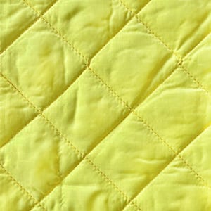 May include: Close-up of a bright yellow quilted fabric. The fabric has a diamond pattern created by stitched lines. The material appears soft and slightly shiny, with subtle variations in the yellow hue across the surface.