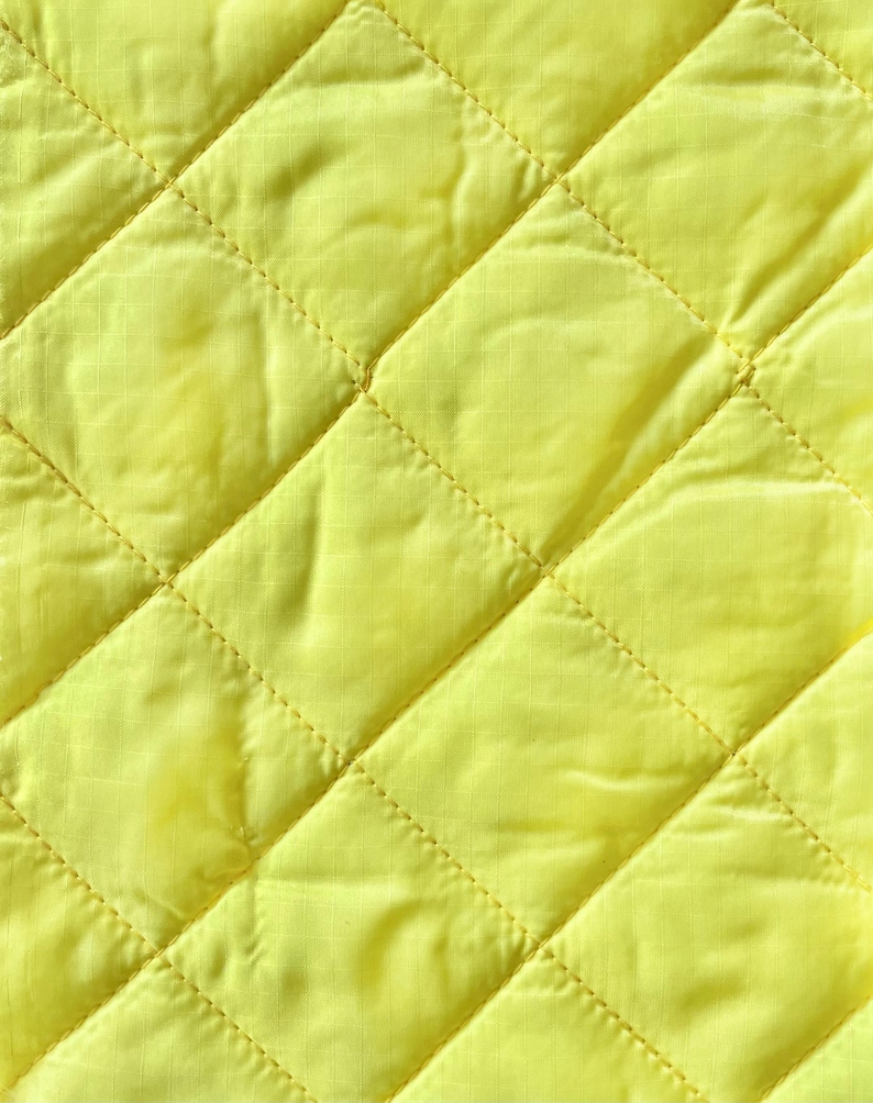 May include: Close-up of a bright yellow quilted fabric. The fabric has a diamond pattern created by stitched lines. The material appears soft and slightly shiny, with subtle variations in the yellow hue across the surface.