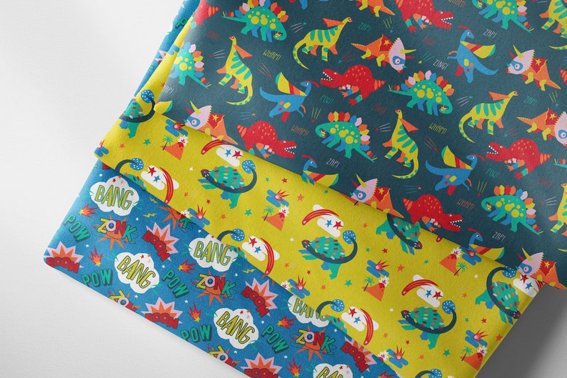May include: Three fabric swatches with different patterns. The top swatch is dark blue with a repeating pattern of colorful dinosaurs and the words "ZOMP", "WHOMP", and "ZING". The middle swatch is yellow with a repeating pattern of colorful dinosaurs and the words "BANG" and "ZONK". The bottom swatch is blue with a repeating pattern of colorful explosions and the words "BANG", "ZONK", and "POW".