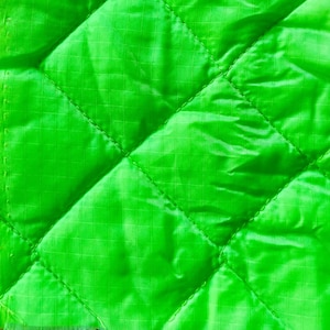 May include: Close-up of a vibrant green quilted fabric. The fabric features a diamond pattern formed by stitched lines. The material has a subtle sheen and a grid-like texture across its surface. The fabric's edges are visible on the left side.